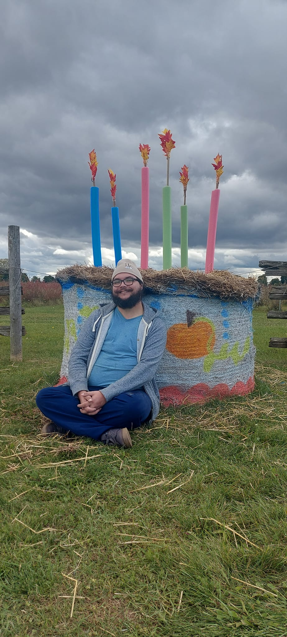 A man with short hair and bushy facial hair sits on the ground in front of a hay bale that has been decorated like a birthday cake, it has six candles made of pool noodles topped with autumn leaves arranged to look like fire